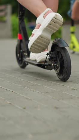 primer plano de una mujer montando un scooter negro en una pasarela pavimentada, sus piernas son visibles a medida que se mueve hacia adelante, con un niño montado en un scooter verde en el fondo rápidamente pasar su