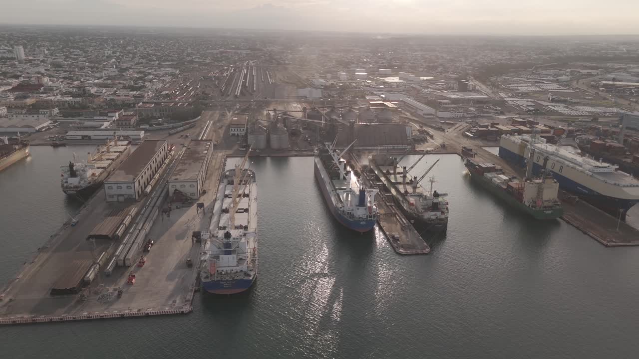 Aerial view of an active port terminal with multiple container and cargo ships docked along the pier. Cranes, storage facilities, and rail lines are visible in the background.