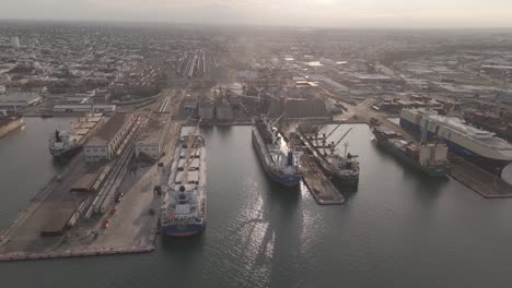 aerial view of an active port terminal with multiple container and cargo ships docked along the pier. cranes, storage facilities, and rail lines are visible in the background.