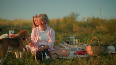 woman feeding dog on leash with watermelon during outdoor picnic, smiling while second woman sits behind, also smiling warmly, dragonfly flying in the background