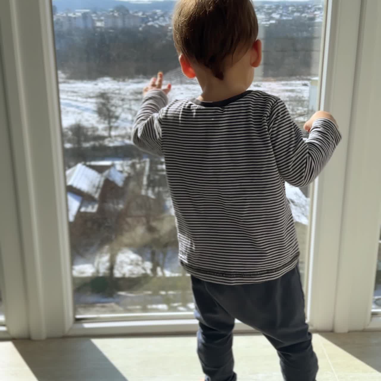 Adorable little baby boy stands near the window on sunny day. Curious toddler slaps the glass with his hand. Vertical screen