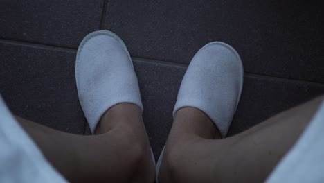 Man-sitting-at-a-spa,-dressing-with-a-robe-and-slippers,-wiggling-his-feet-on-the-dark-black-grey-tile-floor