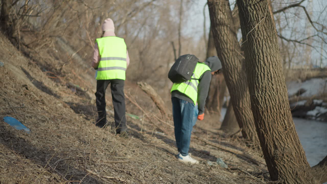 Two volunteers wearing reflective safety vests walking along riverbank during outdoor cleanup, carrying equipment and searching for litter in dry natural area to support environmental conservation