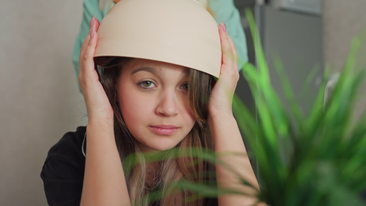 young woman seated in cosy kitchen balancing bowl on head while friend taps bowl with spoon playfully causing laughter and joyful moment next to decorative plant and snack bag