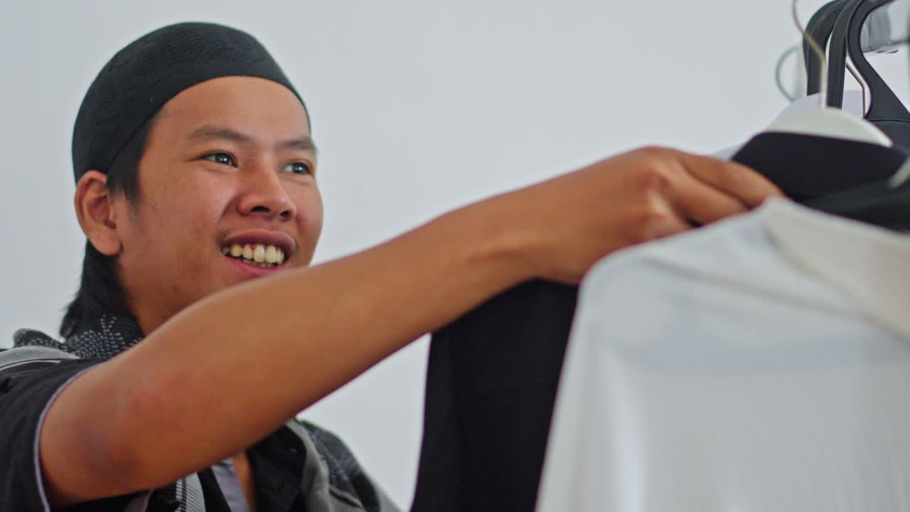 Young Asian Muslim man smiling while choosing clothes from rack for Eid celebration