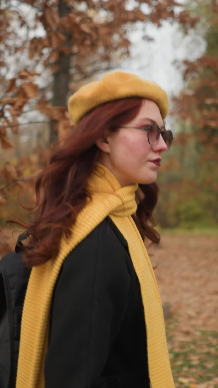 Donna con sciarpa gialla e basco cammina attraverso la foresta, guarda su e giù mentre il vento le soffia i capelli rossi all'indietro, circondata da foglie autunnali.