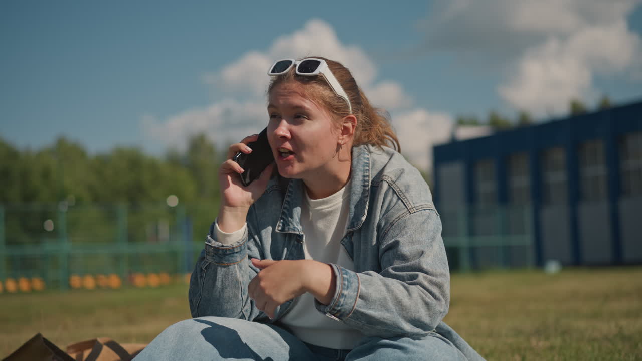 Donna che chiacchiera all'aperto. Studentessa che si gode una giornata di sole. Donna caucasica in giacca di jeans che telefona all'esterno. Giovane donna che si rilassa sull'erba sotto il cielo blu mentre parla al telefono