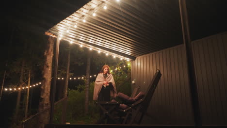 woman sits down in armchair on balcony. rest and tranquility close to nature in rented house for visitors. courtyard illuminated with bright lamps surrounded by dense forest at night