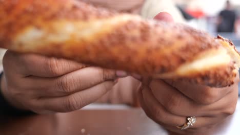 a woman breaks a piece of bread, which looks like turkish simit, with her hands. it has sesame seeds on top and the bread is very golden brown.