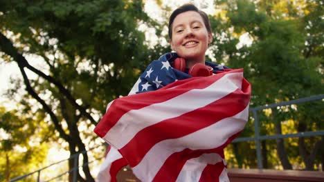 Portrait-of-a-young-girl-in-a-purple-top-and-red-headphones,-who-wraps-herself-with-the-US-flag-in-the-park-in-summer.-A-young-patriot-girl-looks-at-the-camera-and-smiles