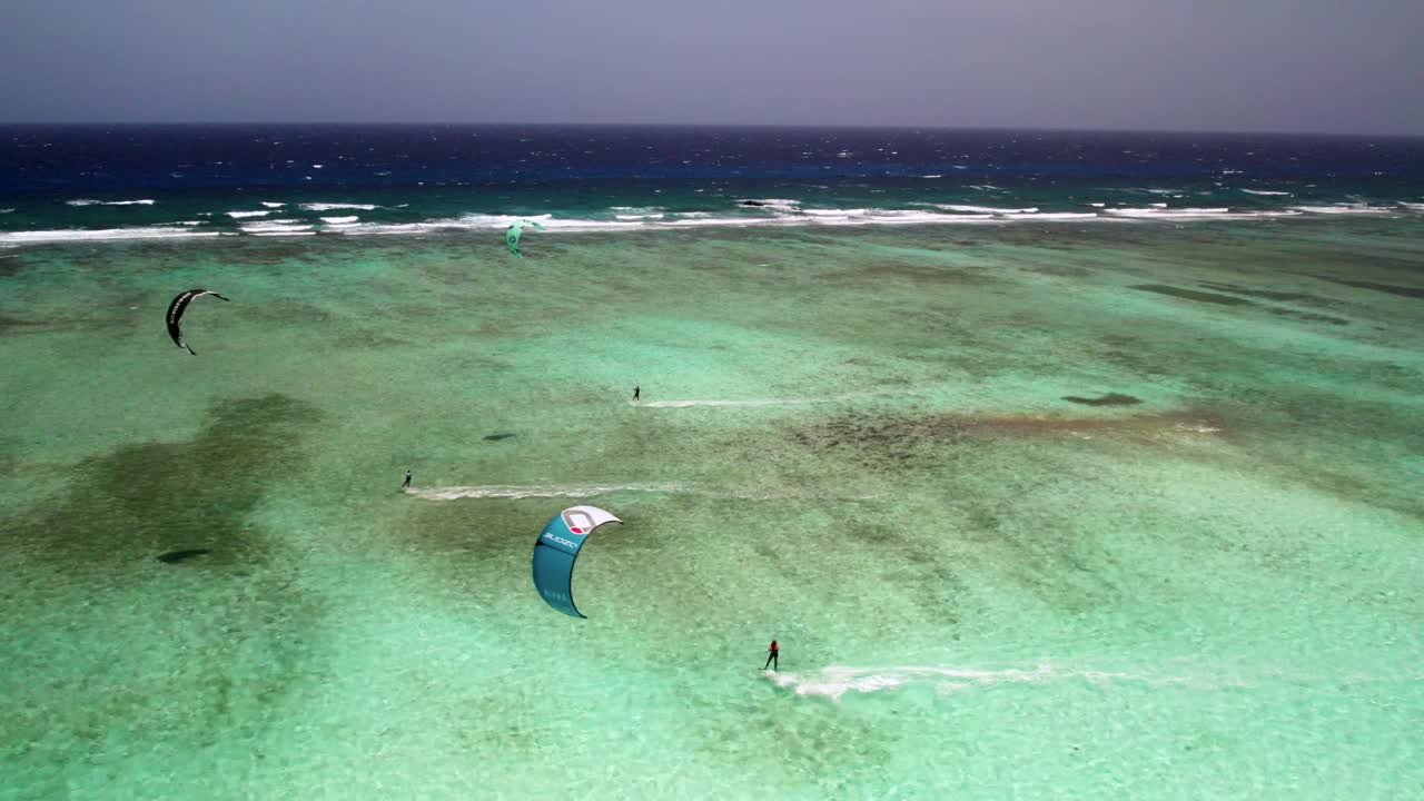 kitesurfistas deslizándose sobre aguas claras de color turquesa junto a una barrera de arrecifes, escena vibrante y activa, día soleado, vista aérea