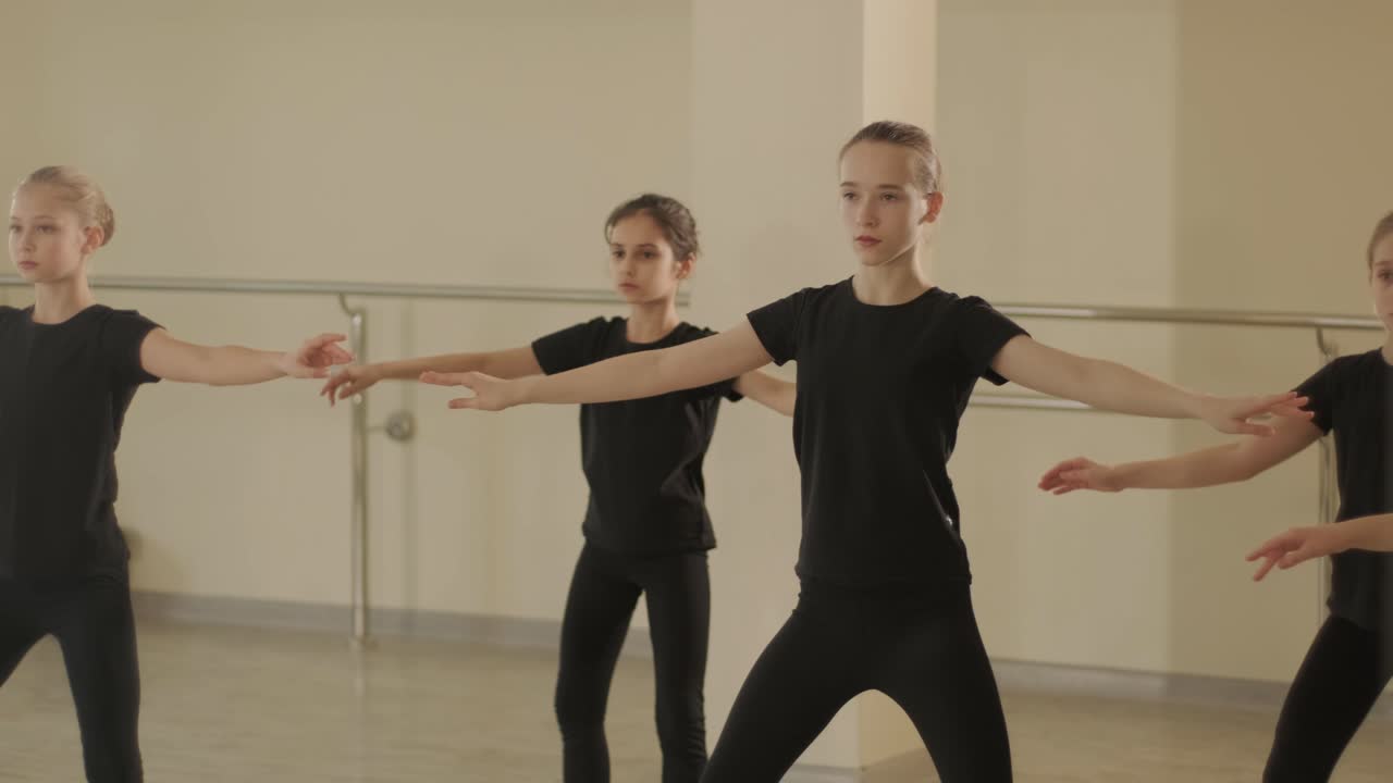 Skupina mladých studentů baletu v černém tanečním oblečení procvičuje pozice v prostorném baletním studiu s dřevěnou podlahou a nástěnnými tyčemi. Soustředěné výrazy a synchronizované pohyby