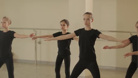 a group of young ballet students in black dancewear practicing positions in a spacious ballet studio with wooden flooring and wall-mounted barres. focused expressions and synchronized movements.