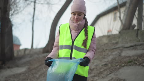 young woman in reflective safety vest and winter clothing preparing blue trash bag during outdoor park cleanup, wearing gloves and beanie, participating in environmental conservation in cold weather