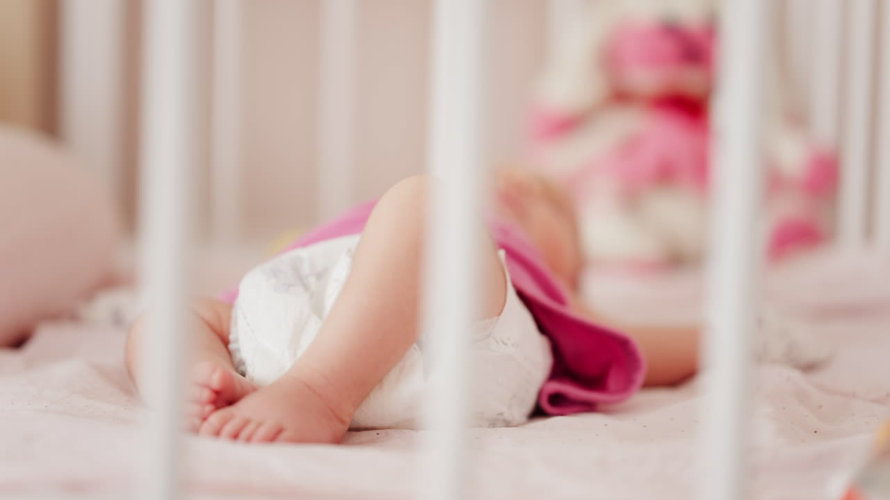 Close up of a baby's legs and diaper as the infant rests in a crib, seen through the crib bars