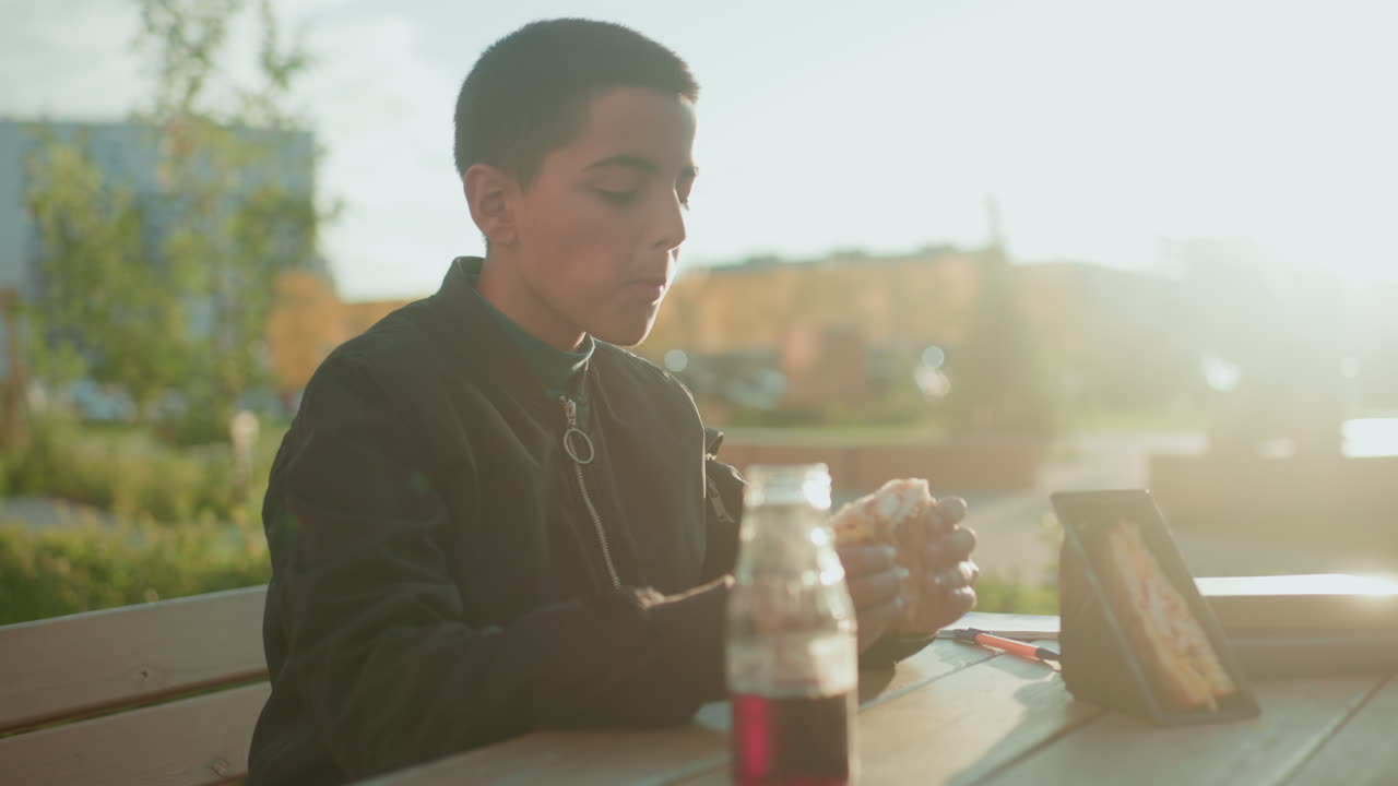 Boy sitting at wooden table outdoors taking bite of sandwich with juice bottle in front and sandwich pack placed nearby under warm daylight in relaxed urban park setting