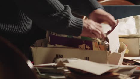 the hands of a person in a blue sweater search through a cardboard box full of old yellowed letters