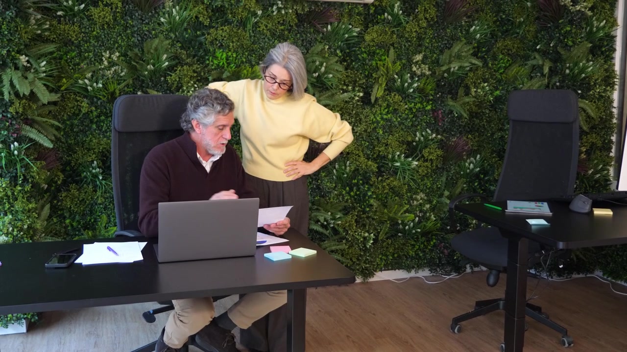 Impiegato senior che consegna un rapporto cartaceo a una collega che si alza dalla sua scrivania per esaminarlo insieme. Concetto di condivisione di documenti, lavoro di squadra, interazione in ufficio e flusso di lavoro.
