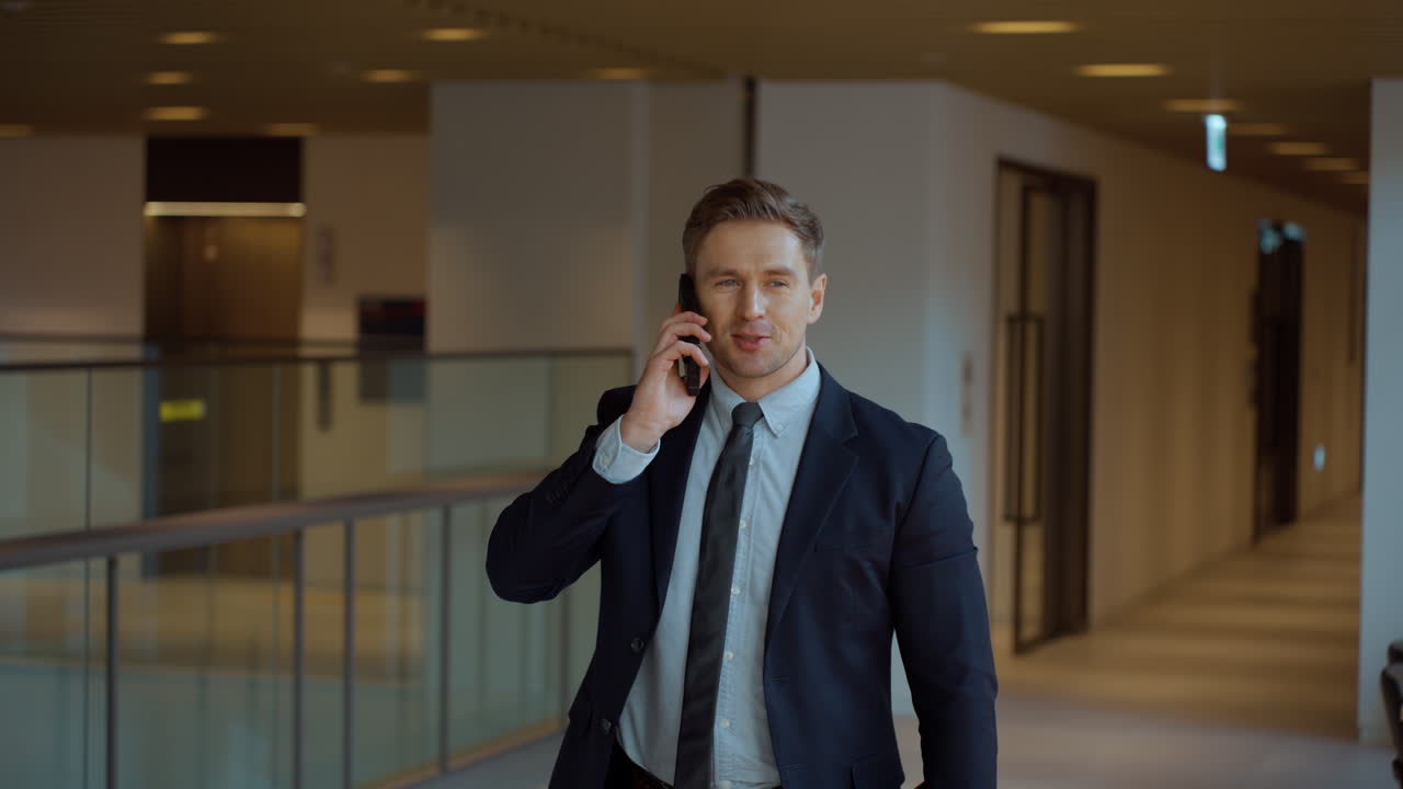Usměvavý pozitivní podnikatel telefonující na smartphonu za chůze dlouhou kancelářskou chodbou – jízda kamery.