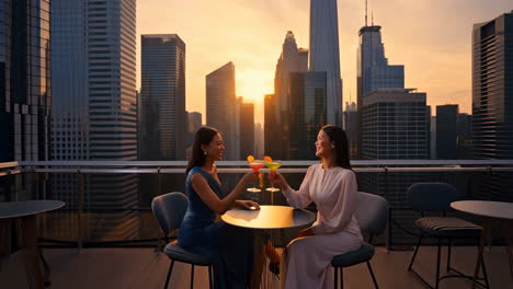 two women celebrating with cocktails on a rooftop bar with a city skyline view at sunset
