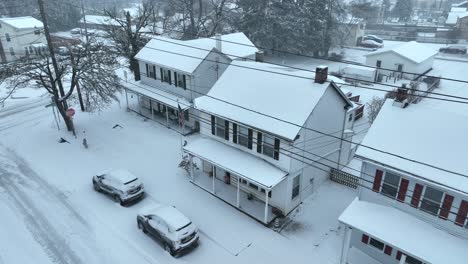 Snowy-street-with-white-houses,-red-accents,-cars,-and-bare-trees