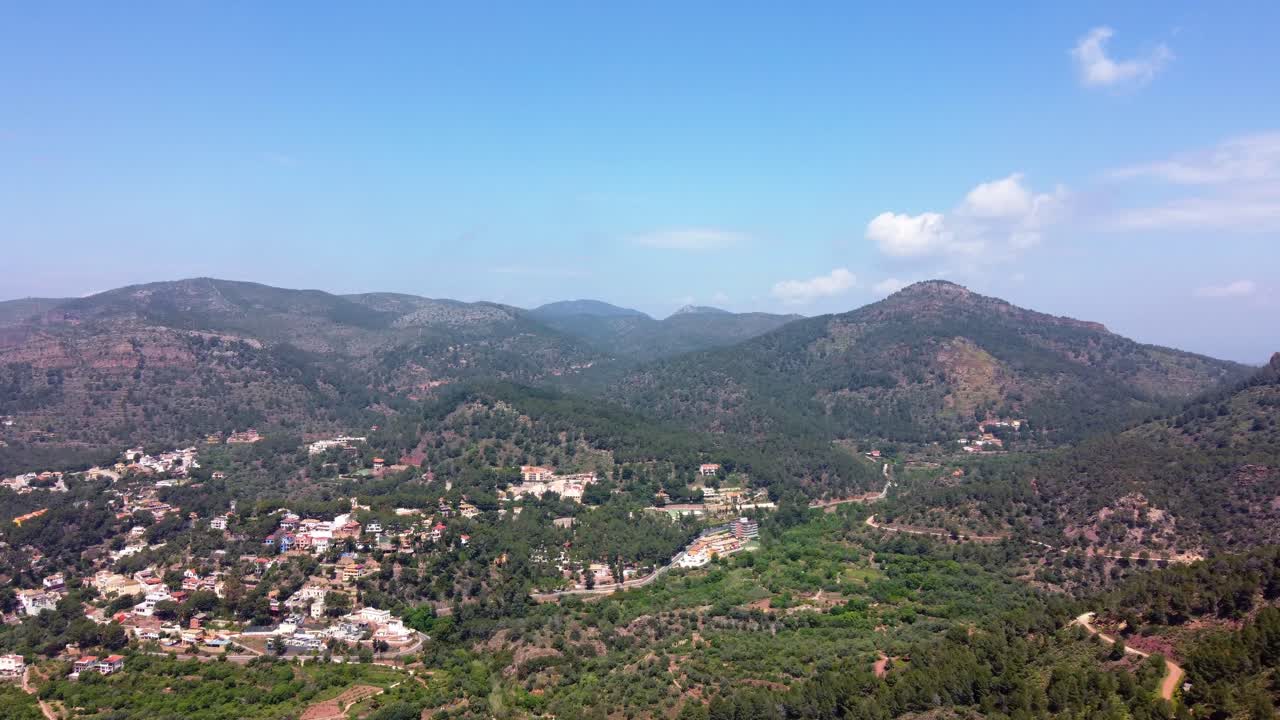Wide 4K aerial view of a Mediterranean mountain landscape in Valencia, Spain, with scattered houses nestled among dense pine forests under a bright blue sky with light cloud coverage