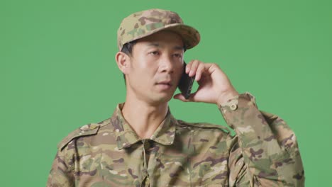 primer plano de un soldado asiático hablando en su teléfono inteligente y mirando a su alrededor mientras camina en el estudio de fondo de la pantalla verde