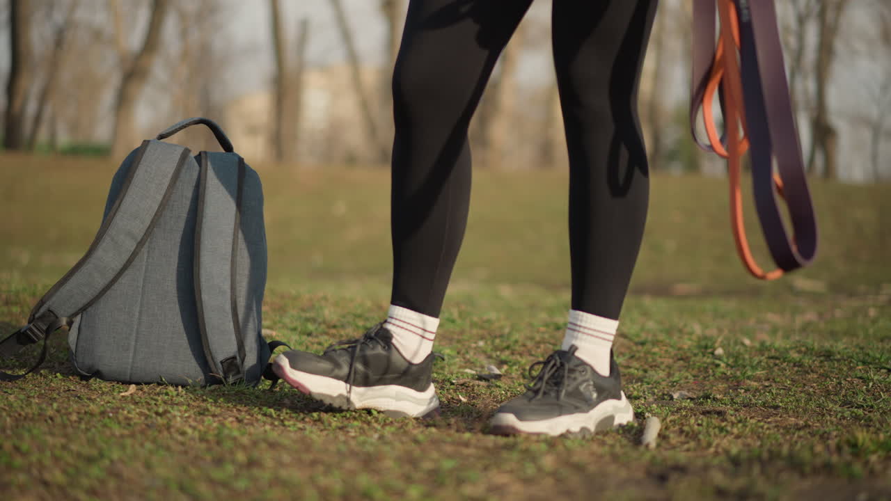 Asian Woman Walking In Park Energetic Stride With Backpack And Colorful Leashes Swinging, Leggings And Running Shoes Show Readiness, Open Field And Bare Trees Emphasize Movement And Active Lifestyle