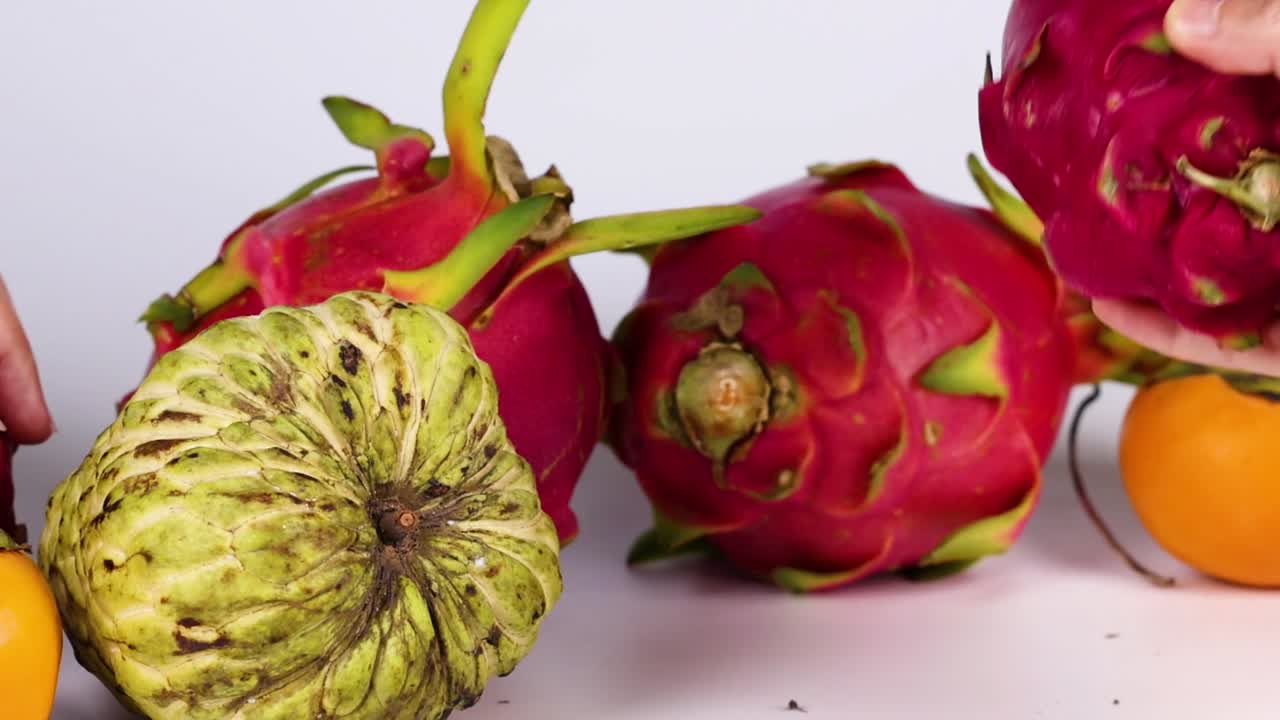 Hands selecting dragon fruits and cherimoya from a vibrant arrangement with an orange.