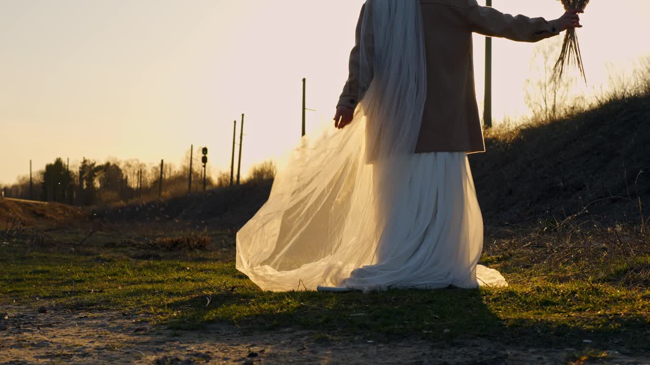 A bride in soft sunset light turns gracefully, holding grass, wearing a white dress and beige jacket.