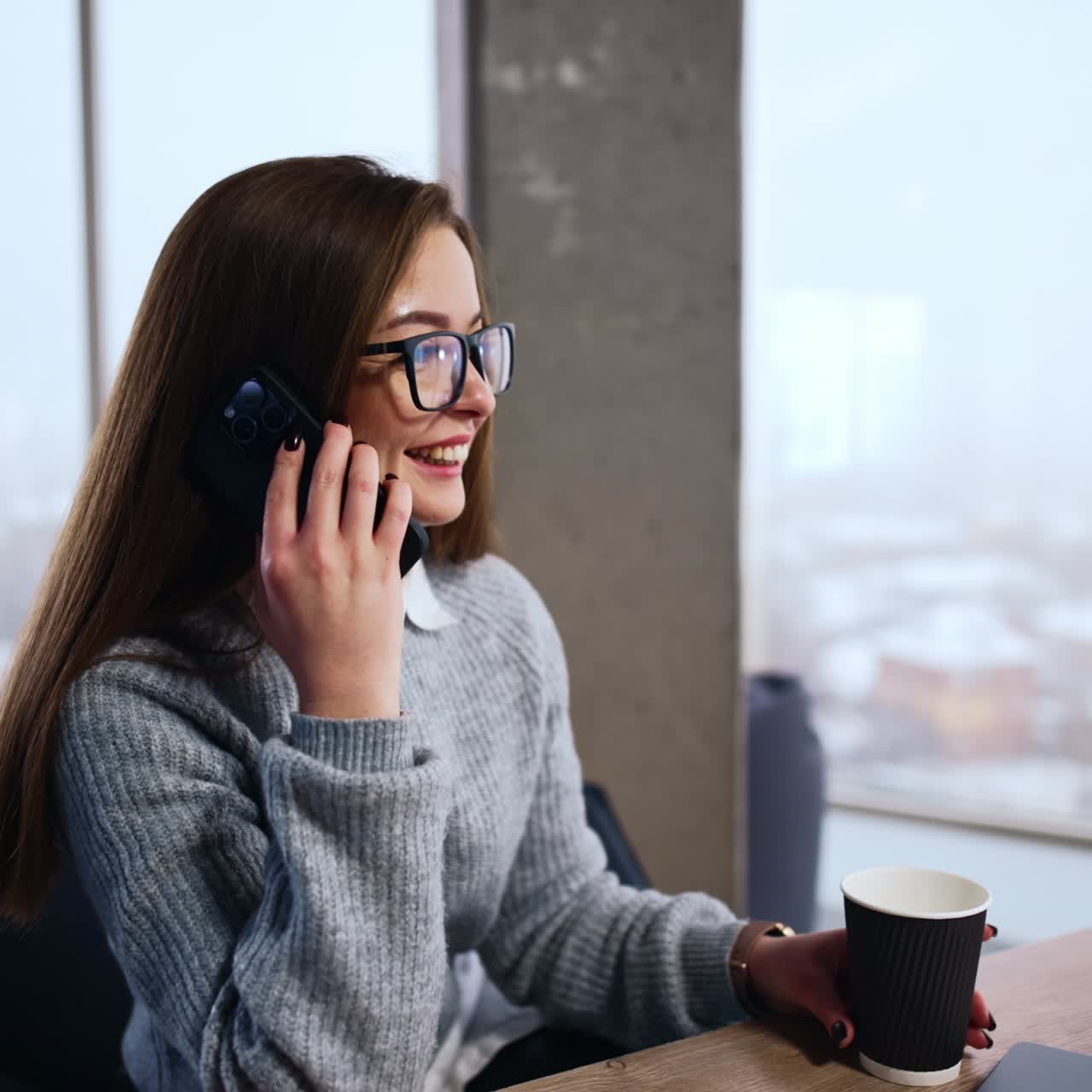 Una donna con gli occhiali siede alla scrivania parlando al suo smartphone. La signora che tiene un bicchiere di carta ride mentre parla al telefono.
