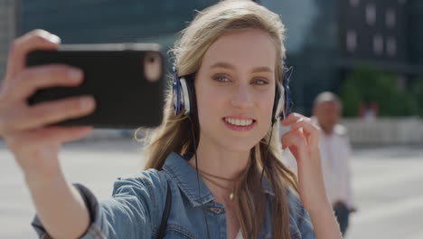 portrait cute young blonde woman using smartphone taking selfie photo in city enjoying listening to music wearing headphones on sunny urban day