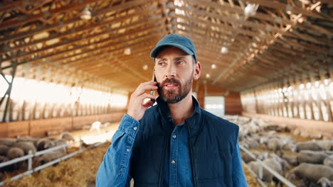 close-up view of young caucasian man in cap walking in stable with sheep flock and talking on mobile phone