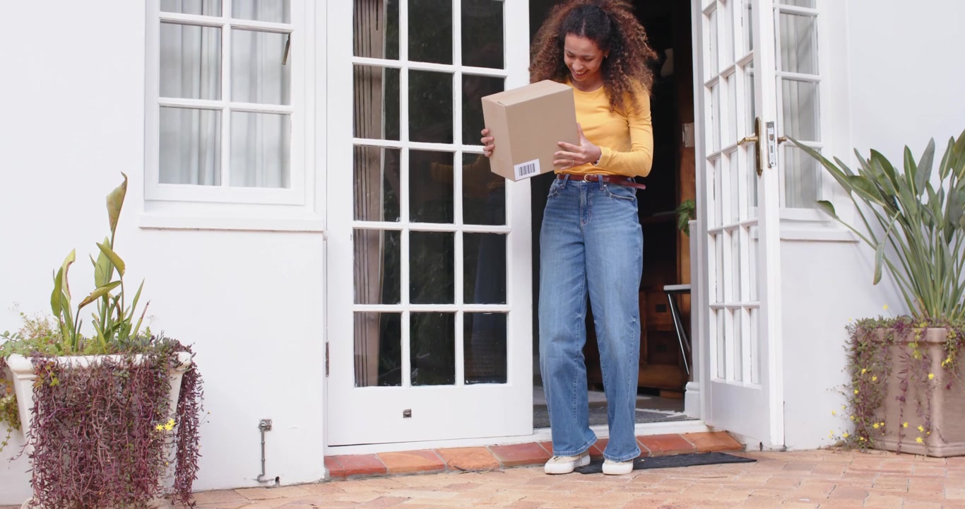 Adult woman in mustard top noticing package, opening white doors, stepping onto patio, lifting box