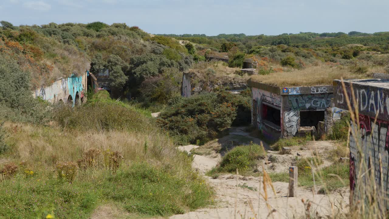 Wide shot of graffiti-covered concrete bunkers, overgrown vegetation, sandy dunes, bright daylight, static camera