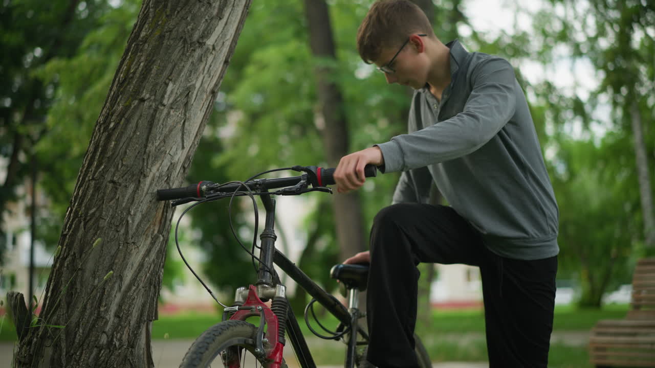 un niño se acerca a una bicicleta estacionada cerca de un árbol en un campo cubierto de hierba, colocando el pie en el pedal mientras se centra en ajustar algo, en el fondo, otro niño se aleja