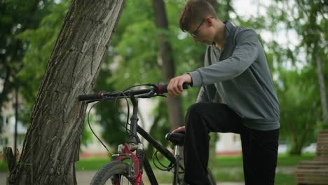 young boy approaches a parked bicycle near tree in a grassy field, placing foot on the pedal while focused on adjusting something, in the background, another boy is walking away