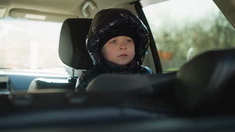 a close-up of a child seated inside a car, wearing a shiny black jacket and a black beanie, gazing thoughtfully out the window