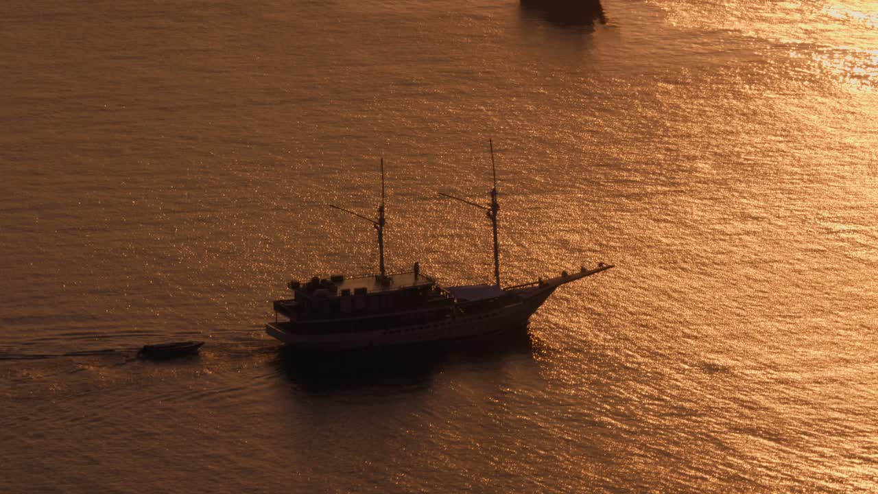 Aerial view of a sailboat gracefully sailing across golden waters against a backdrop of a setting sun