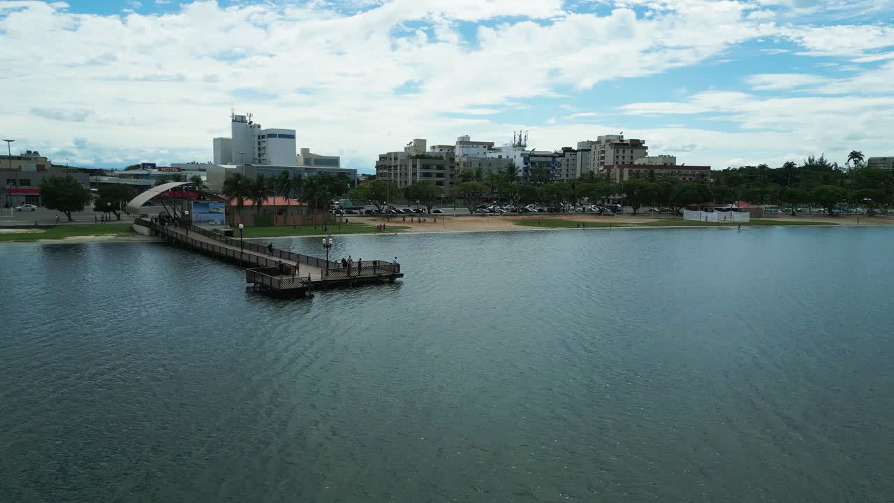 Aerial orbital drone shot captures the wooden pier and the dock in Araruama' shore providing a view of the people walking along the boardwalk. As the drone orbits, it reveals the city architecture.