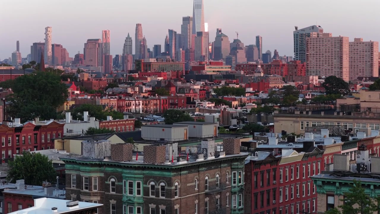 Historiske brownstone-bygninger med udsmykkede gesimser fylder forgrunden i Hoboken, New Jersey, tætte rækkehuse i mursten strækker sig mod en diset Manhattan-skyline, der gløder ved solnedgang. Bredt dronebillede fra luften
