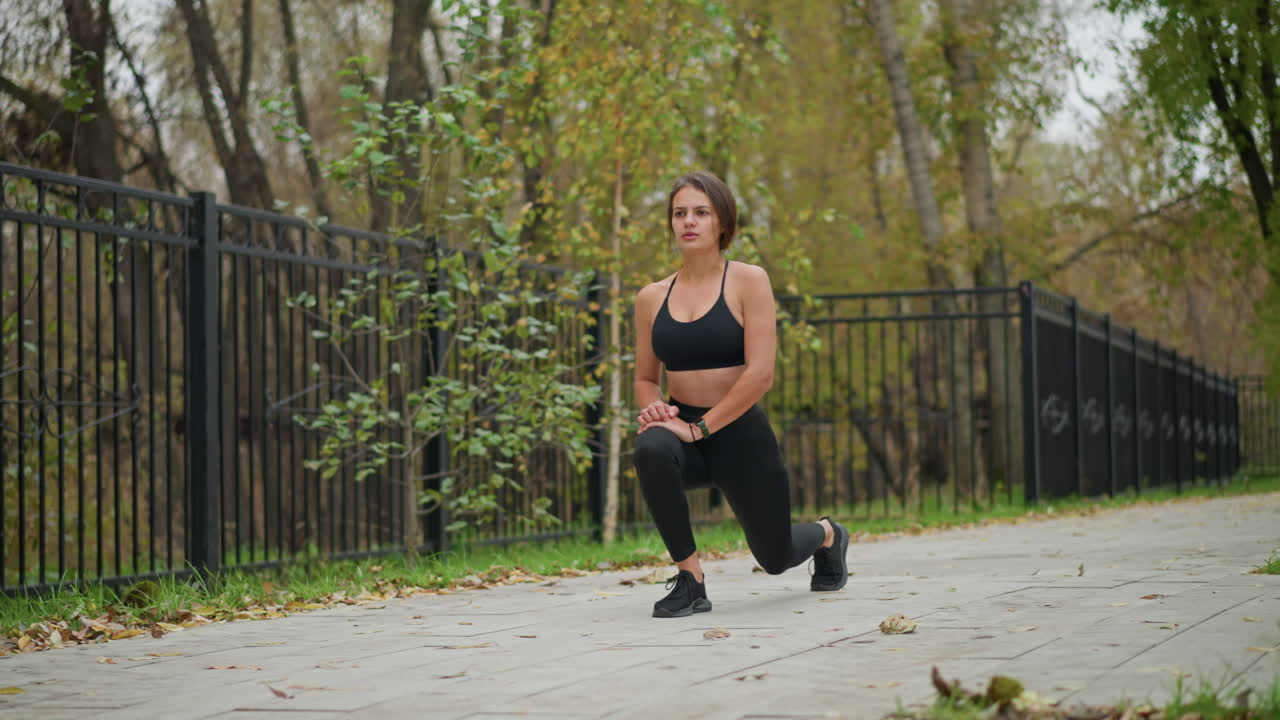 Sporty woman in black fitness wear performing leg stretch outdoors in a park, background shows iron fence and trees as she exercises, focusing on flexibility and body conditioning