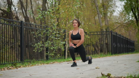 sporty woman in black fitness wear performing leg stretch outdoors in a park, background shows iron fence and trees as she exercises, focusing on flexibility and body conditioning