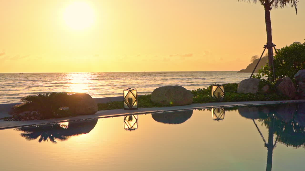 Summertime: beautiful view of the sunset at the pool area with palm, stones, and night lamps in the foreground. Sunlight reflections in pool water