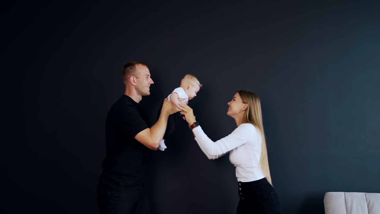 Playing with an infant baby. Adorable kid is waved by his dad and mom clapping hands to her kid. Black backdrop.
