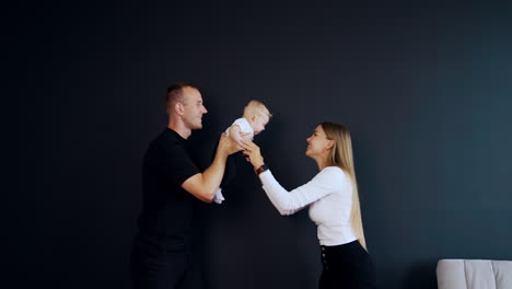 playing with an infant baby. adorable kid is waved by his dad and mom clapping hands to her kid. black backdrop.