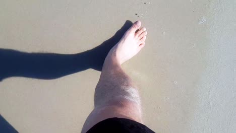 top view of man's feet walking on sandy beach barefoot splashed by sea waves