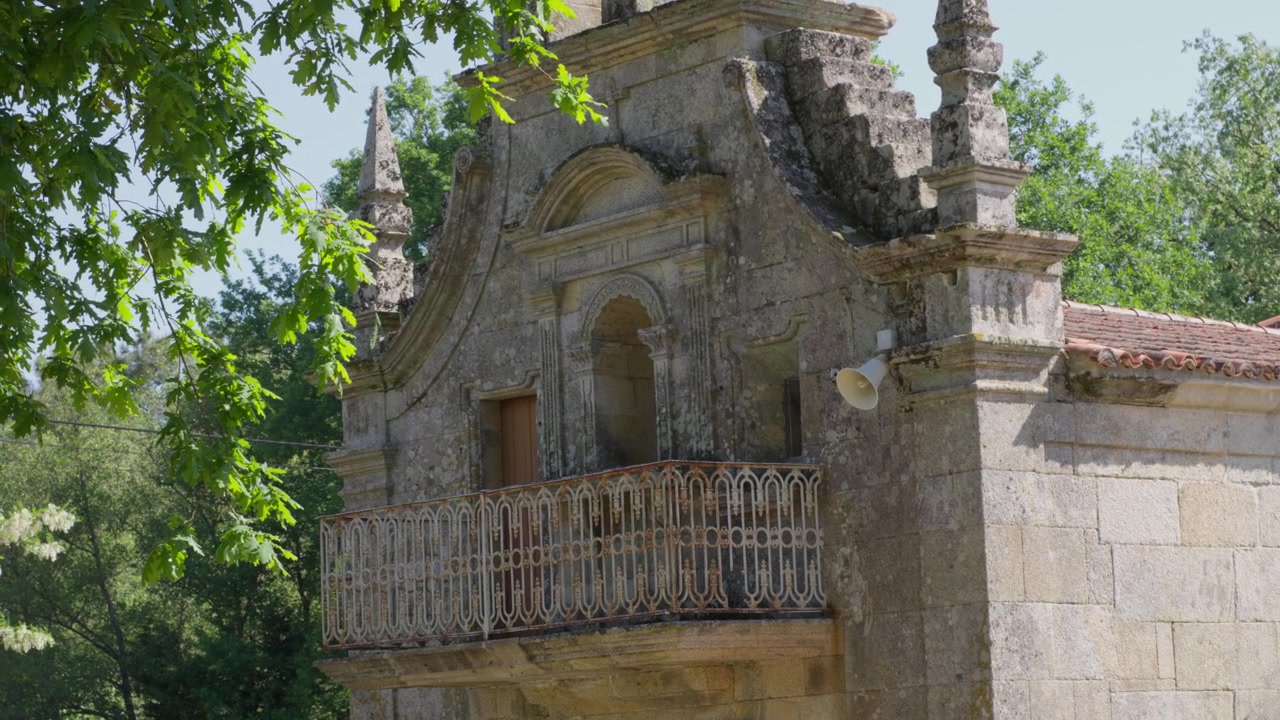 Facciata di una cappella in pietra ornata con balcone a balaustra, dettagli architettonici e vegetazione lussureggiante, catturata nella calda luce naturale del giorno. Nessuna persona presente.
