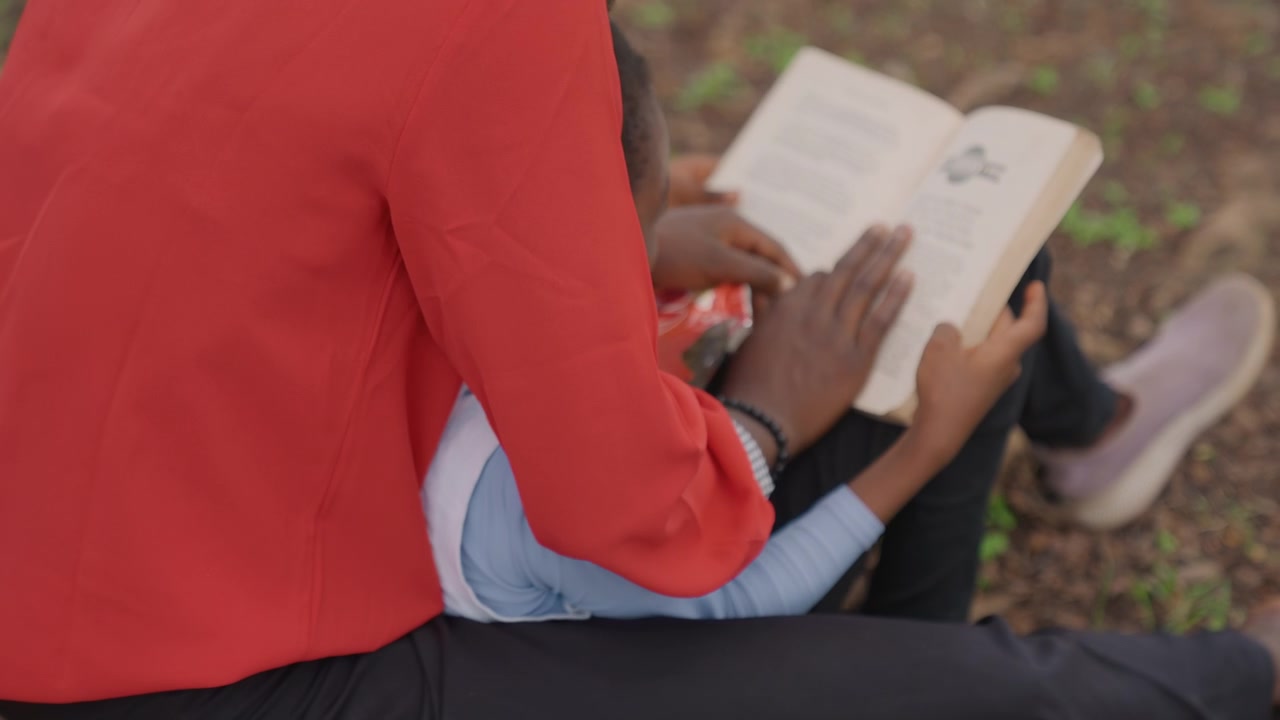 Mãe e filha negras em aula de leitura ao ar livre, mãos guiando as páginas, close-up do livro e dedo apontando, blusa vermelha e pulseira visíveis, atmosfera de aprendizado focada, instrução paciente e prática de alfabetização