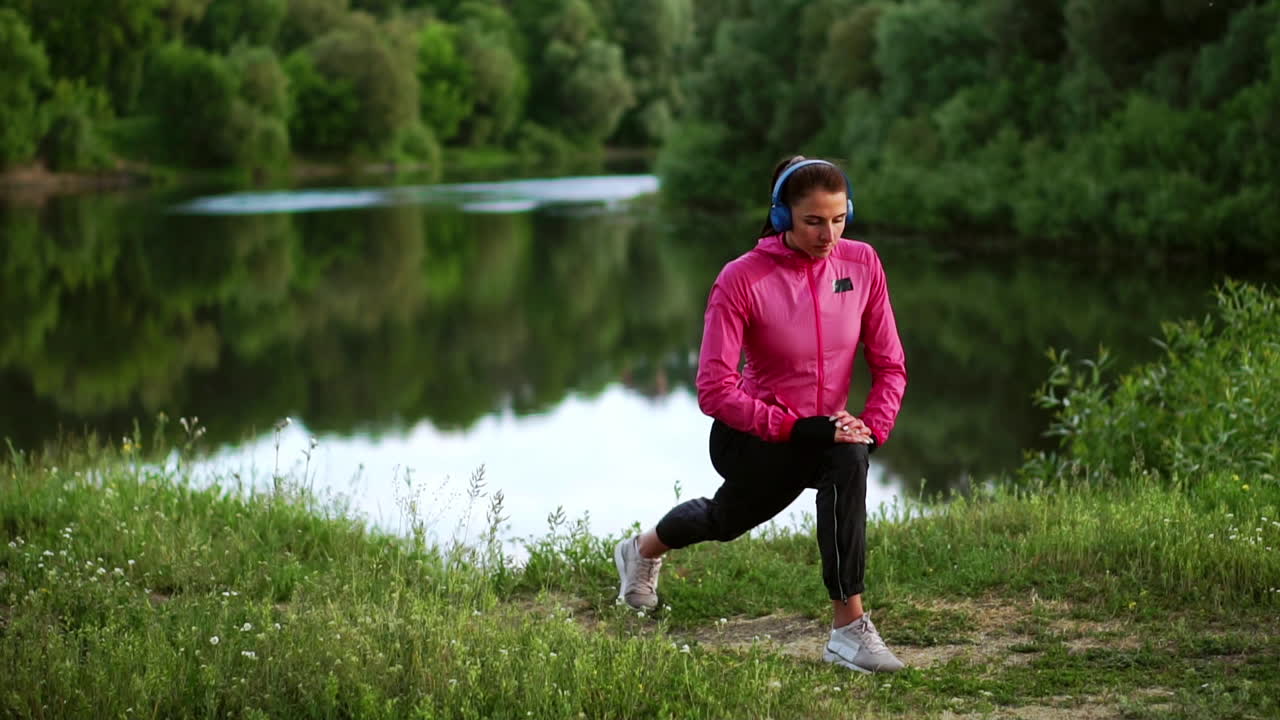 A girl in a pink jacket is preparing for a run warm up and listen to music in headphones through the phone